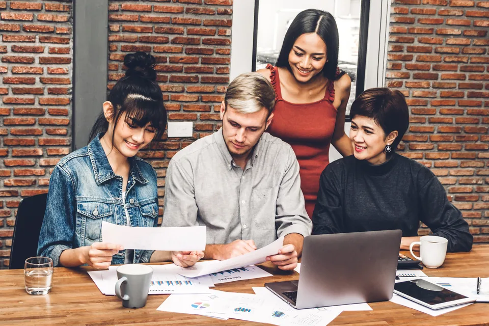 Four colleagues review printed charts together beside an open laptop in a brick-walled office.