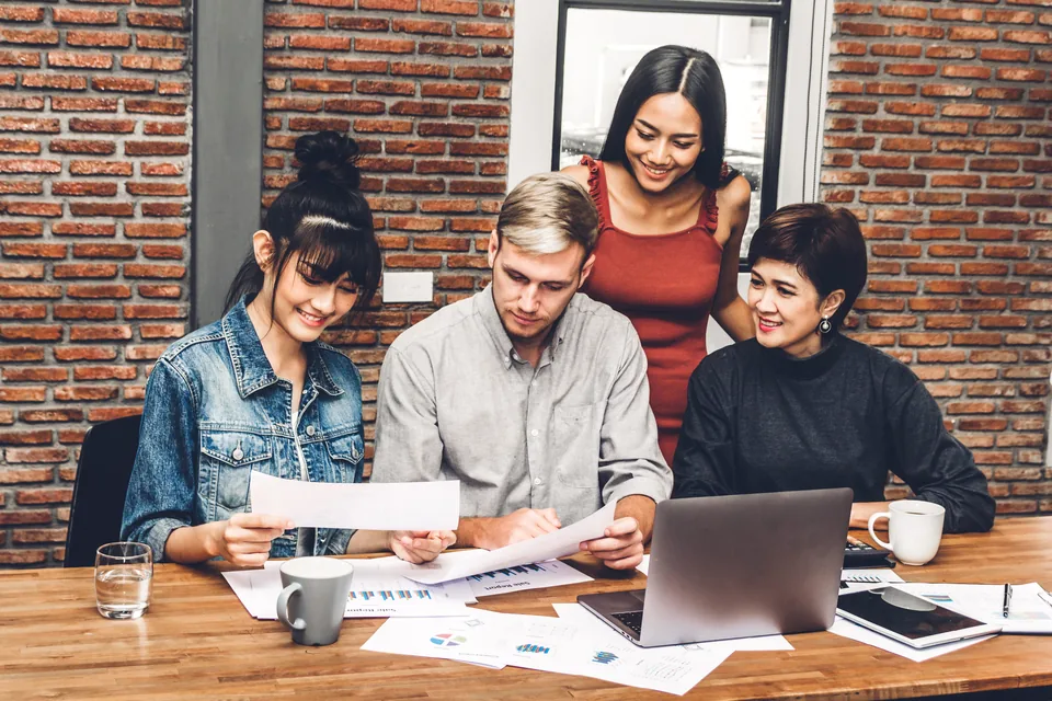 Four colleagues review printed charts together beside an open laptop in a brick-walled office.