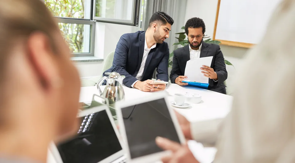Colleagues at a workshop table with laptops, tablets, and printed notes.